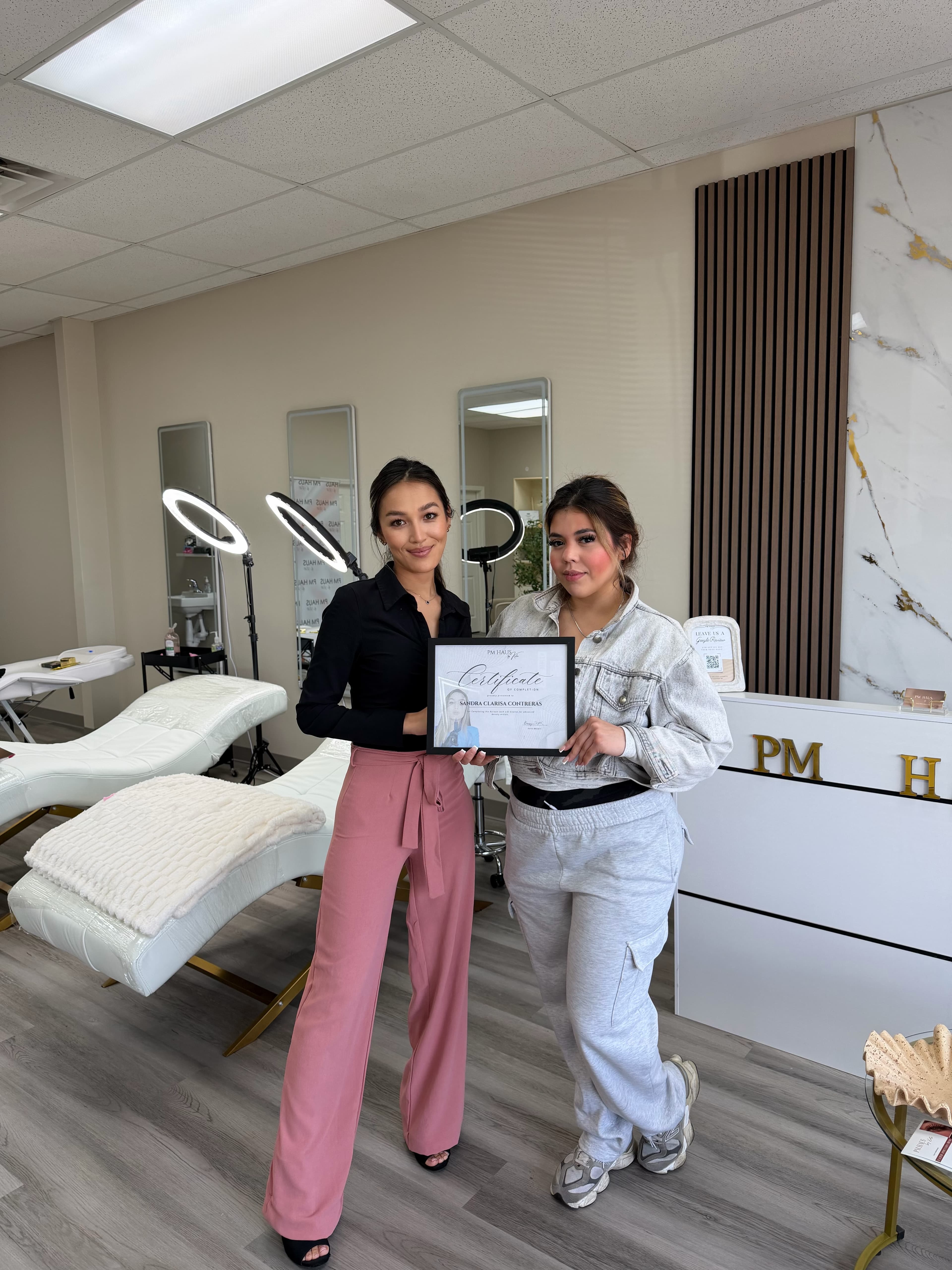Two women posing with a framed certificate in a bright, modern beauty salon training studio.